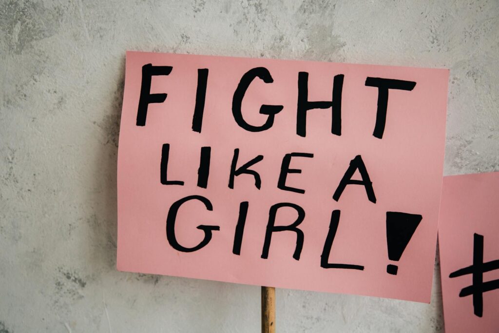 Close-up of a 'Fight Like a Girl' message on a pink placard against a gray wall.